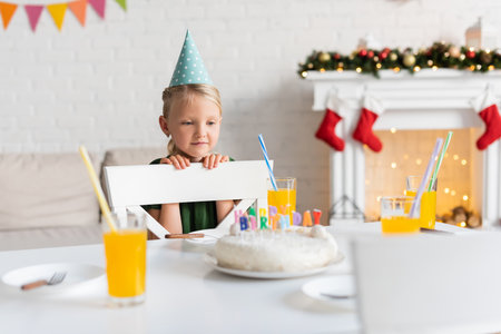 Kid in party cap looking at glasses with orange juice and birthday cake at homeの写真素材