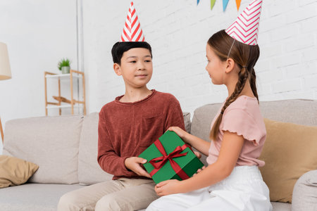 Asian boy in party cap giving present to friend during birthday party at homeの写真素材