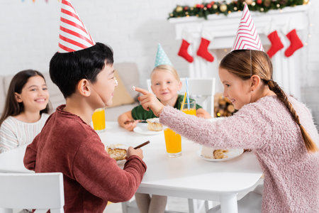 Kid holding cake cream on finger near asian friend during birthday celebration at homeの写真素材