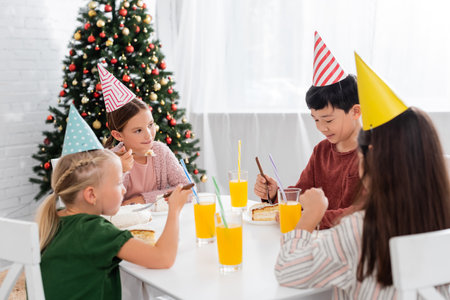 Multiethnic kids in party caps celebrating birthday near cake and orange juice at homeの写真素材