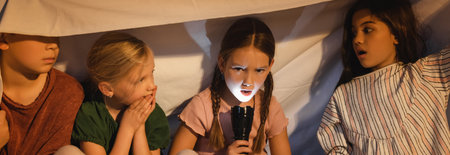 Girl holding flashlight near frightened friends under blanket at home, bannerの写真素材