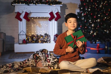 Asian boy holding gift box while sitting near blurred christmas tree at homeの写真素材