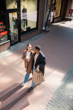 high angle view of interracial couple in trendy coats walking with shopping bags and coffee to go along showcasesの素材 [FY310192566437]