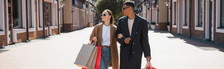 smiling multiethnic couple in trendy coats and sunglasses carrying shopping bags, bannerの写真素材