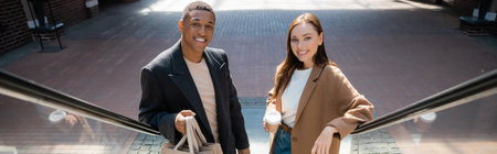 trendy multiethnic couple with shopping bags and coffee to go smiling at camera on escalator, bannerの写真素材