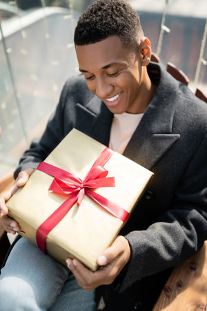 happy african american man in coat sitting with gift box outdoorsの写真素材