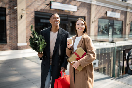 young woman with coffee to go and gift box smiling near stylish african american boyfriend with small christmas treeの写真素材