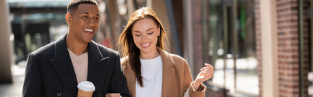 happy african american man with takeaway drink walking with stylish girlfriend on city street, bannerの写真素材