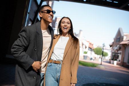 cheerful woman in trendy coat laughing near african american man in sunglasses on street in cityの写真素材