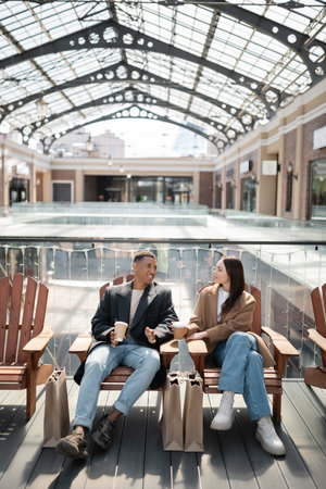 trendy african american man talking to girlfriend while sitting near shopping bags and blurred mallの写真素材