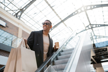 joyful african american man in sunglasses looking away on escalator while holding shopping bags and paper cupの写真素材