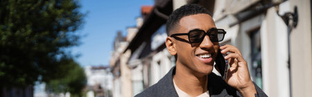 happy african american man in trendy sunglasses talking on smartphone on blurred street, banner