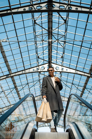 low angle view of happy and stylish african american man with shopping bags and takeaway drink on escalator under transparent roofの写真素材