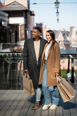 full length of multiethnic couple in fashionable clothes standing with shopping bags on blurred city backgroundの写真素材