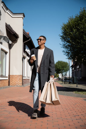 full length of fashionable african american man in sunglasses walking with shopping bags and paper cup on streetの写真素材