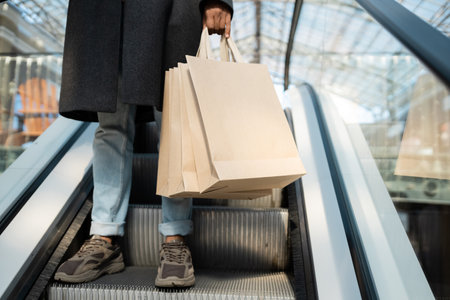 cropped view of african american man in coat and sneakers holding shopping bags on escalatorの写真素材