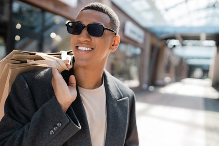 happy african american man in coat and sunglasses holding shopping bags on blurred streetの写真素材