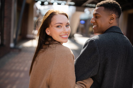 pretty and happy woman looking at camera near african american boyfriend on blurred streetの写真素材