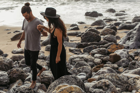 bearded man holding hands with girlfriend in dress and hat while walking on rocks near ocean