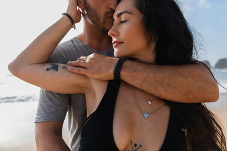 portrait of bearded man kissing tattooed girlfriend with closed eyes near oceanの写真素材