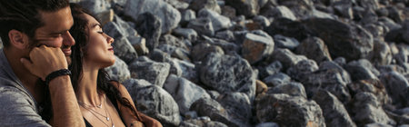 bearded man hugging tattooed woman with closed eyes near rocks on beach, banner