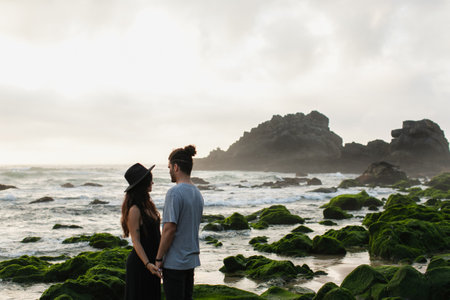 woman in hat and dress holding hands with bearded boyfriend near mossy stones and oceanの写真素材