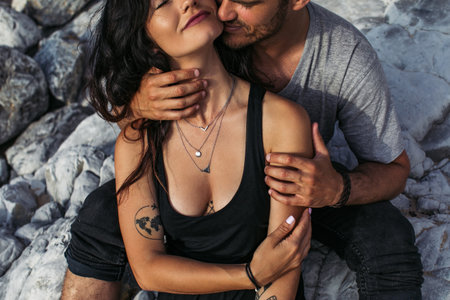cropped view of bearded man kissing tattooed woman near rocks on beachの写真素材