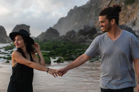 pleased woman in hat and dress holding hands with bearded boyfriend near ocean