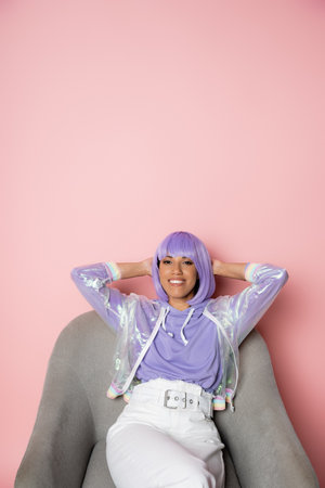 joyful african american woman in purple wig sitting on grey armchair on pinkの写真素材