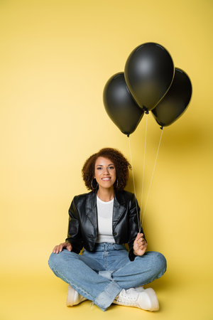 full length of pleased african american woman in leather jacket and jeans sitting with black balloons on yellowの写真素材