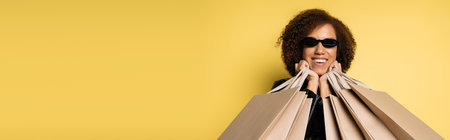 cheerful african american woman in sunglasses holding shopping bags on black friday on yellow, bannerの写真素材