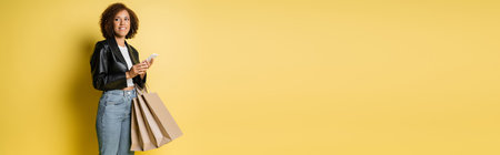 pleased african american woman in stylish leather jacket holding shopping bags and mobile phone on yellow, bannerの写真素材