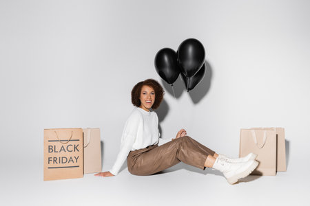 cheerful african american woman in autumnal outfit holding black balloons near shopping bags on greyの写真素材