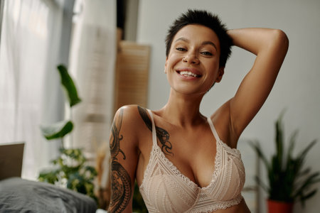 happy tattooed woman in beige bra holding hand behind head and smiling at cameraの写真素材