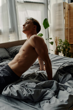 side view of young and sexy man sitting on grey bedding at homeの写真素材