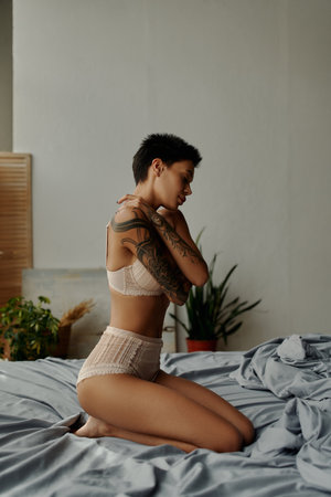 side view of seductive tattooed woman in lingerie embracing herself while sitting on bedの写真素材
