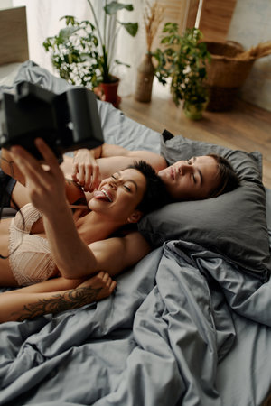 Sexy woman in bra taking selfie on vintage camera near young boyfriend on bedの写真素材