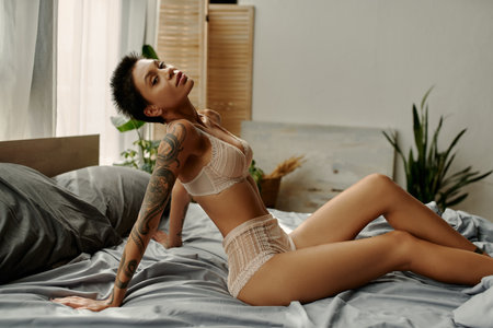 Tattooed woman in beige underwear looking at camera on bedの写真素材
