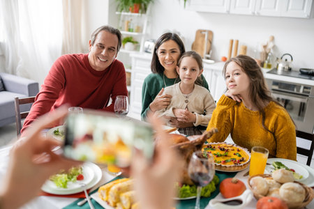 Interracial family smiling near delicious thanksgiving dinner and woman taking photo on smartphone at homeの写真素材