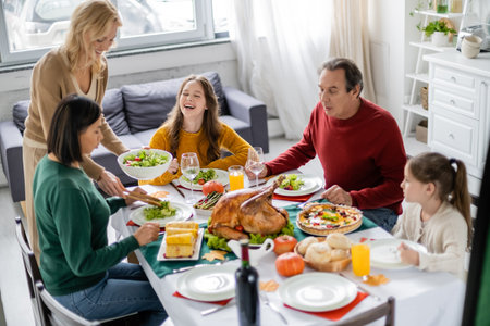 Cheerful teenager sitting near multiethnic family and tasty thanksgiving dinner at homeの写真素材