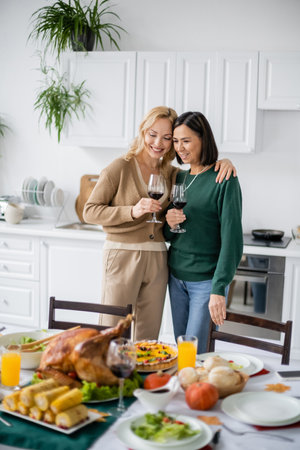 Cheerful woman holding glass of wine and hugging multicultural mom during thanksgiving at homeの写真素材