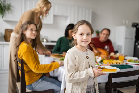 Girl holding tasty pie near blurred family celebrating thanksgiving dinner at homeの写真素材