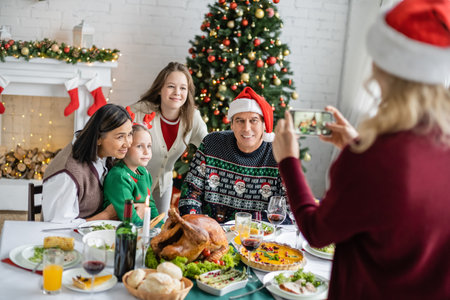 blurred woman taking photo of happy interracial family near festive dinner and christmas treeの写真素材