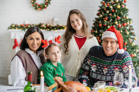 cheerful girls with multiracial grandparents looking at camera near blurred festive dinner and decorated christmas treeの写真素材