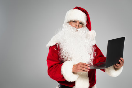 Santa claus in eyeglasses and costume holding laptop isolated on greyの写真素材