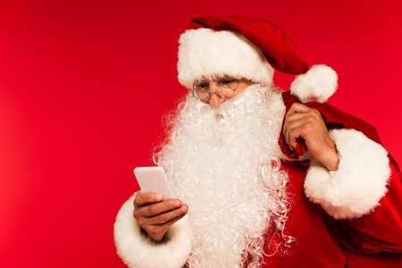 Santa claus in eyeglasses holding sack and using smartphone isolated on redの写真素材