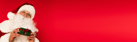 Santa claus in eyeglasses holding small gift isolated on red with copy space, bannerの写真素材