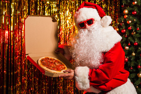Santa claus in sunglasses holding takeaway pizza near christmas tree and tinselの写真素材