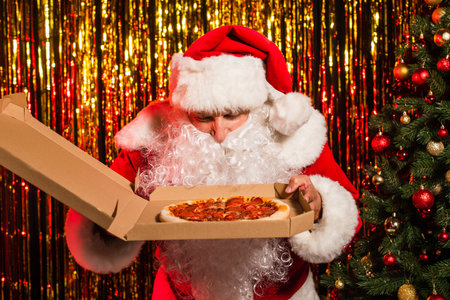 Santa claus in hat holding takeaway pizza near christmas tree and tinselの写真素材
