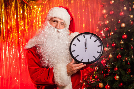 Santa claus in costume holding clock near christmas tree and tinsel